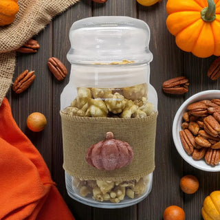 Glass jar with popcorn and a burlap sleeve featuring a pumpkin design, surrounded by pumpkins and pecans on a wooden surface.
