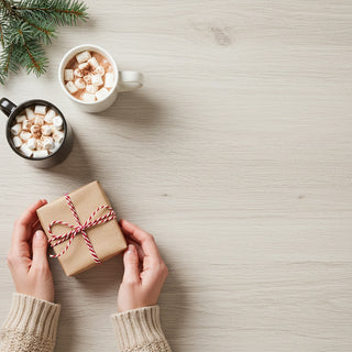 Two mugs of hot chocolate with marshmallows, a wrapped gift, and Christmas tree branches on a light wooden surface.