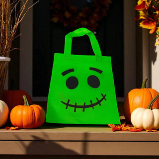 Green Halloween treat bag with a face design on a table with pumpkins.