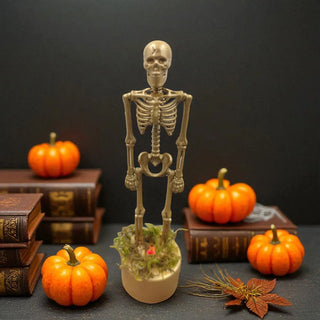 Skeleton figure with a potted plant surrounded by pumpkins and books on a dark background