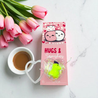 Pink packaging of 'Hugs & Squishies' with a green toy, next to pink tulips and a cup of coffee on a marble surface.