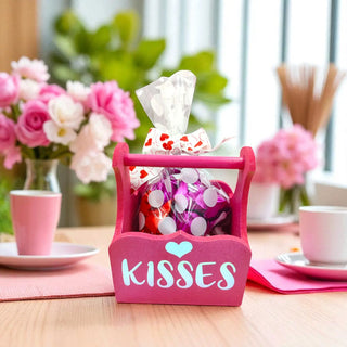 Pink candy holder with 'Kisses' text on a table with flowers and cups in the background