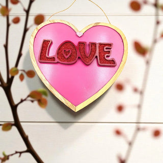 Heart-shaped pink ornament with 'LOVE' in red glitter on a wooden background