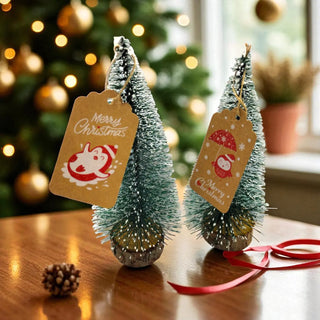 Two small Christmas trees with 'Merry Christmas' tags on a wooden surface, with a blurred Christmas tree in the background.
