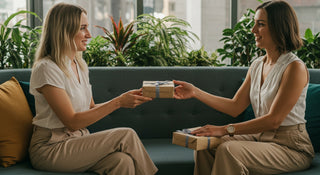 Two women sitting on a couch exchanging gift boxes in an indoor setting with plants in the background.
