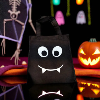 Black Halloween tote bag with a face design on a reflective surface with Halloween decorations in the background.