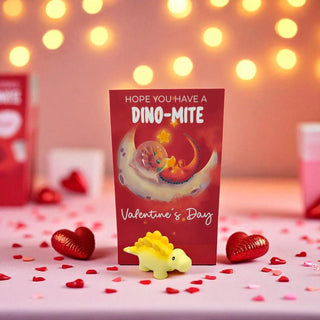 Valentine's Day card with dinosaur illustration and small yellow dinosaur toy on a decorative surface with heart-shaped confetti and lights in the background.
