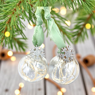 Two clear glass ornaments with snowflake designs hanging from a green ribbon on a Christmas tree.