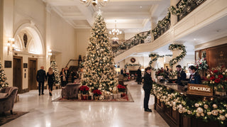 A large, beautifully decorated Christmas tree in a grand four-star hotel lobby with marble floors, a staircase and a concierge desk styled with holiday greenery and flowers.