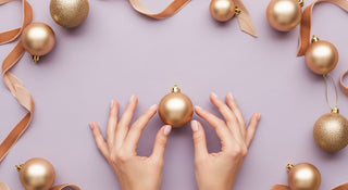 Gold Christmas ornaments and ribbons on a light purple background with hands reaching for an ornament.