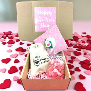 Valentine's Day gift box with heart-shaped treats, a lollipop, and a small bag on a pink background with heart decorations.