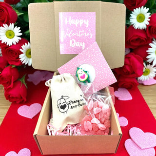 Valentine's Day gift box with a lollipop, small bag, and pink heart-shaped treats on a red background with flowers.