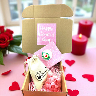 Valentine's Day gift box with a card, small bag, and heart-shaped items on a pink surface with red roses and candles.