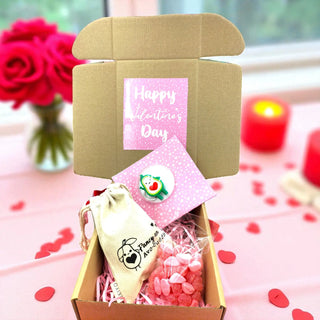 Valentine's Day gift box with a card, small bag, and decorative items on a pink surface with flowers and candles in the background.