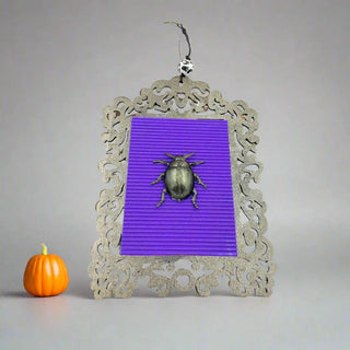 Decorative frame with purple background and metallic beetle, next to a small pumpkin on a gray background