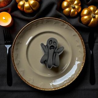 Beige plate with a ghost decoration on a dark tablecloth with gold pumpkins and a candle.