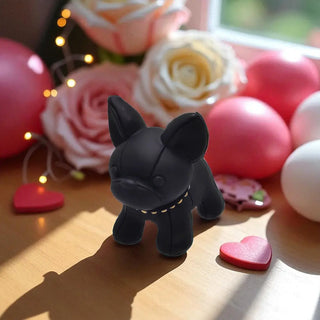 Black dog figurine on a wooden surface with pink and white flowers and balloons in the background.