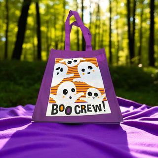 Purple tote bag with Halloween design featuring ghosts and 'BOO CREW!' text, placed on a purple fabric with a forest background.
