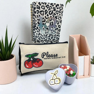 Pouch with cherry design and text, leopard print notebook, and small container on a light surface.