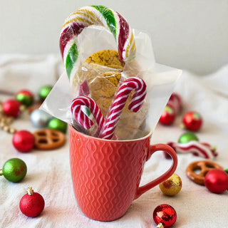 Red mug with cookies and candy canes surrounded by Christmas ornaments on a light background