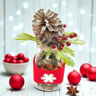 Decorative jar with pine cone, berries, and greenery on a light wooden surface with festive decorations.