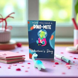 Valentine's Day card with dinosaur illustration on a table with small toys and stationery.