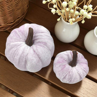 Two light purple pumpkins with brown stems on a wooden surface, with a vase of white flowers in the background.