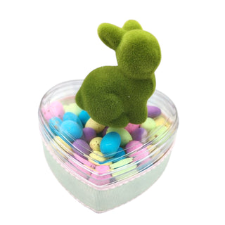 Heart-shaped container with colorful candies and a green moss-like bunny toy on a white background