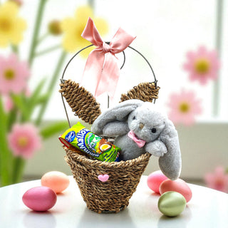 Easter basket with a plush bunny, candy, and eggs on a table with flowers in the background.