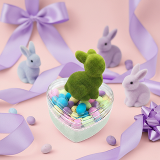 Green moss rabbit in a clear container with pastel balls, surrounded by purple ribbons and small rabbit figurines on a pink background.
