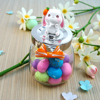 Glass jar with colorful candies and a bunny-shaped lid on a wooden surface with flowers.