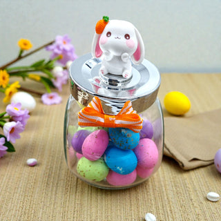 Glass jar with colorful candies and a rabbit-shaped lid on a wooden surface with flowers in the background.