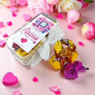 Jar of gold and purple heart-shaped candies with a decorative label on a pink background with flowers and hearts.