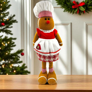 Gingerbread man plush toy in a chef outfit on a wooden surface with Christmas decorations in the background.