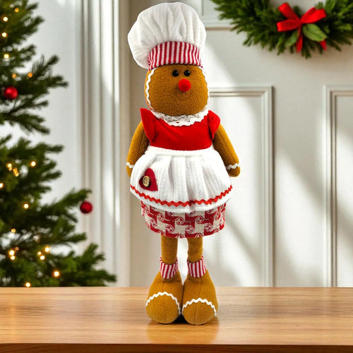 Gingerbread man plush toy in a chef outfit on a wooden surface with Christmas decorations in the background.