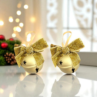 Two gold Christmas ornaments with bows on a reflective surface with blurred lights in the background.
