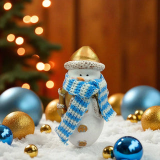 Snowman figurine with a gold hat and blue scarf surrounded by Christmas ornaments on a wooden background.