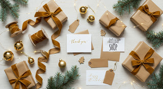 Gift boxes with brown ribbons, Christmas ornaments, and cards on a light wooden surface.