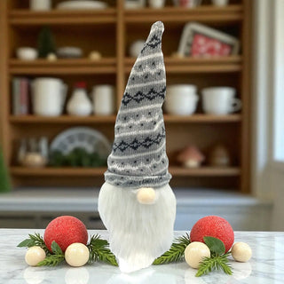 Decorative gnome with a patterned hat on a marble surface with Christmas ornaments, in front of a bookshelf.