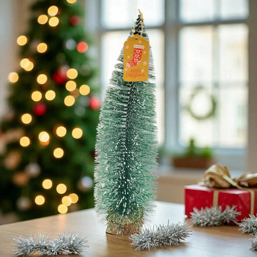 Small decorated Christmas tree with a tag on a table, blurred Christmas tree and gift in the background