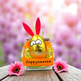 Gold chocolate egg with bunny ears in an orange Easter basket on a wooden surface with pink flowers and a blurred floral background.