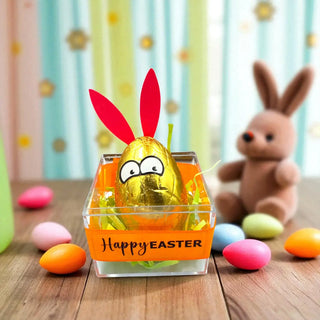 Gold chocolate egg with bunny ears in an 'Happy Easter' box, surrounded by colorful Easter eggs and a toy rabbit.