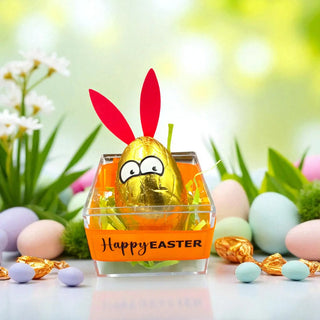 Gold chocolate egg with bunny ears in a clear container labeled 'Happy Easter' on a festive background.