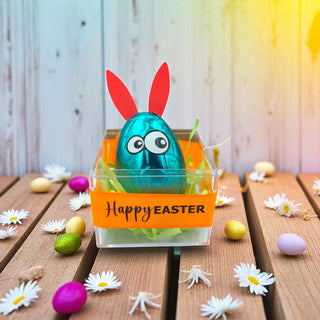 Blue Easter egg with bunny ears in a clear container on a wooden table with Easter decorations.