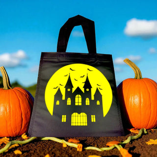 Black Halloween tote bag with a yellow haunted house design, surrounded by pumpkins against a blue sky.