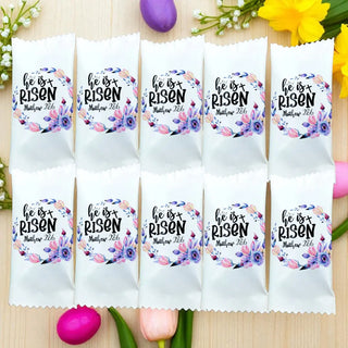 Packets with 'He is Risen' text and floral designs on a wooden surface with Easter eggs and flowers.