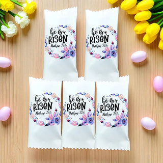 White bags with 'He is Risen' text and floral design on a wooden surface with Easter eggs and tulips.