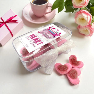 Pink heart-shaped candy in a clear container with a cup of coffee and flowers on a white surface.