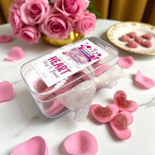 Pink heart-shaped candies in a clear container with a white bow, surrounded by pink roses and candy on a marble surface.