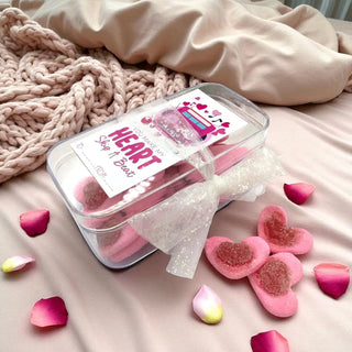 Heart-shaped bath bombs in a clear container with a decorative bow on a soft surface with pink petals.